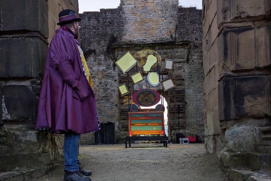 A man dressed in a three quarter length purple dress coat with bowler hat stands at the entrance of a stone walled building. Through the entranceway we can see an elabourate dresser illuminated in the dusky evening