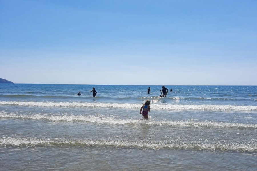 People play in the sea as small waves come onto a sandy beach. The sky is bright blue and it's sunny.