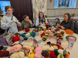 A mum, gandma and two kids sit around a table that is full of coloured wool and pom poms. They all look happy.