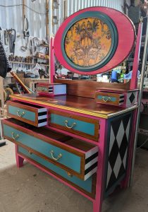 A dressing table stands in a workshop. It has been painted bright colours of pink, blue and yellow