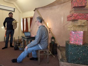 Two men are in a rehearsal room practicing their lnes for the christmas show. One is sat on a stool, the other one is standing up.