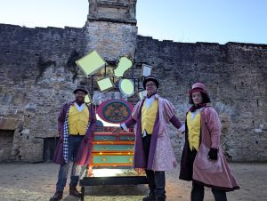 Two men and woman sstand infront of an illuminated dresser. The are wearing colourful costumes and big pink thre quarter length jackets. They are in a walled courtyard and is it starting to get dark.