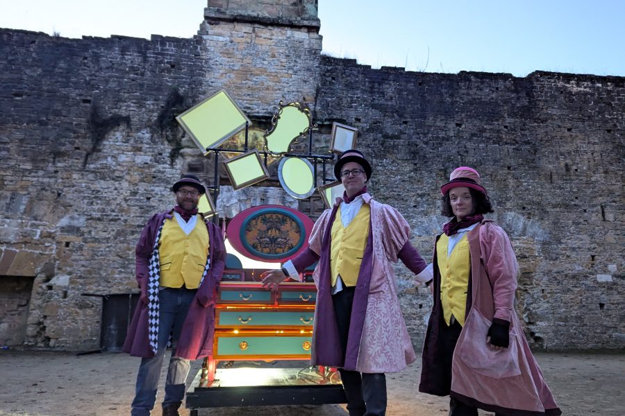 Two men and woman sstand infront of an illuminated dresser. The are wearing colourful costumes and big pink thre quarter length jackets. They are in a walled courtyard and is it starting to get dark.