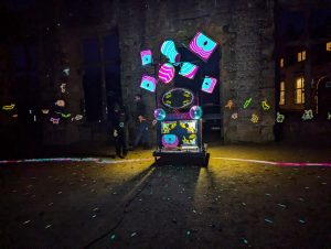 An illuminated dresser stands in an open stone coutyard at night. It is lit up and patterns dance on the walls to create a magical atmosphere