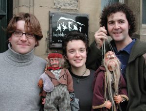 Three young faced people look at the camera with smiling faces holding up puppets.