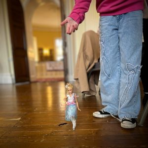 In the photo you can only see the lower section of a girls body. She is wearing blue jeans and a pink jumper holds a barbie on a string like a puppet. She has redesigned Barbie's outfit.