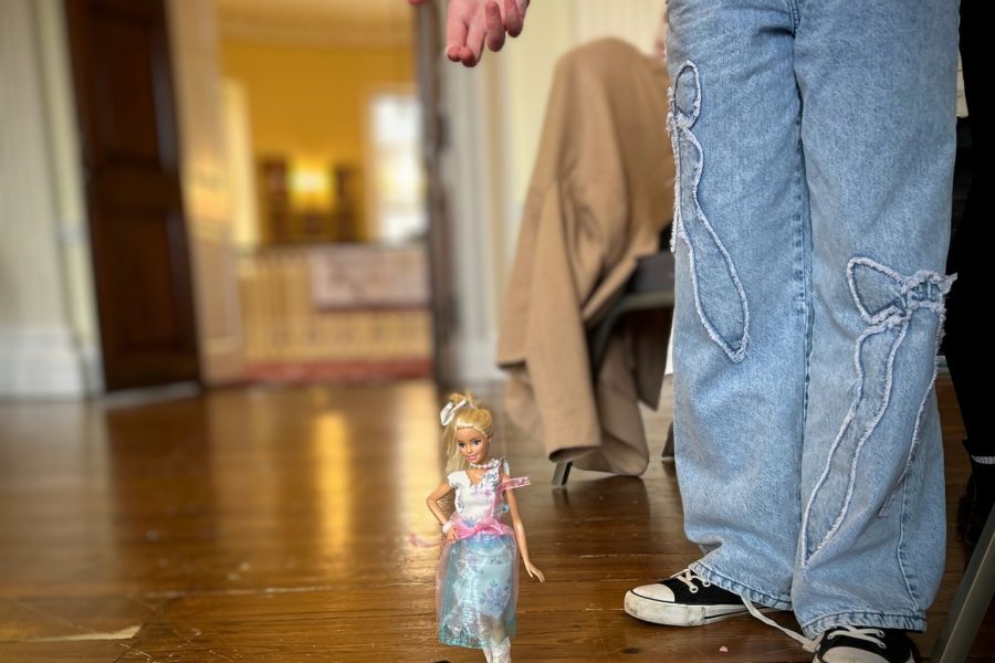 In the photo you can only see the lower section of a girls body. She is wearing blue jeans and a pink jumper holds a barbie on a string like a puppet. She has redesigned Barbie's outfit.