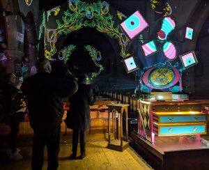 a brightly painted dresser stands in fron of the pews in a church. It is dark and the dresser is illuminated and projected images of tree branches adorn the space. People have their backs to the camera as they view the digital artwork.