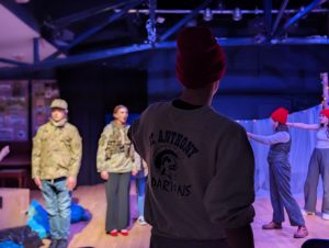A person has their back to the camera and is looking towards a theatre space. They are wearing a red hat and grey jumper with writing on the back.