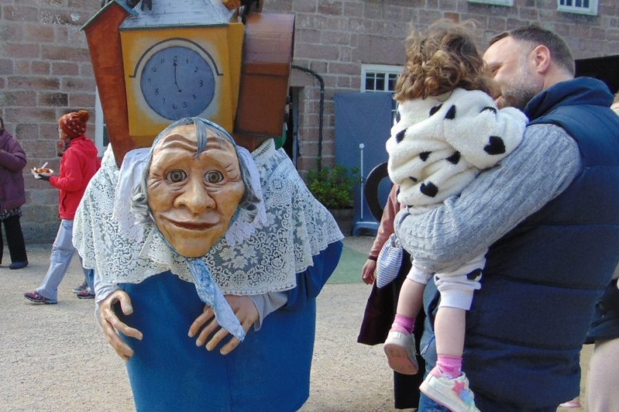 A large walkabout granny puppet stops to look at a child in her dads arms. They are looking at her.