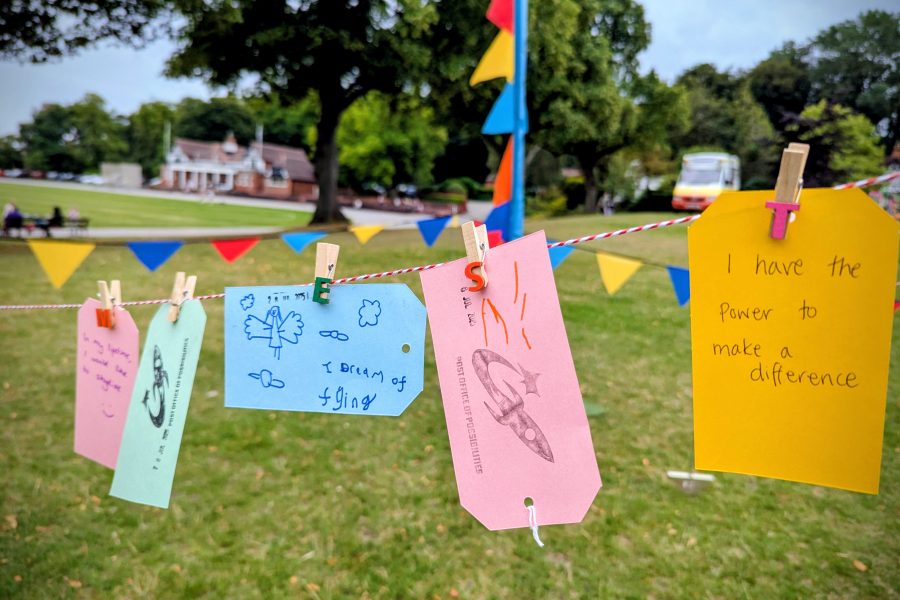 Coloured labels pegged onto a line. The background is a green park with grass and treees and there is coloured flags too.