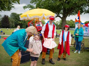 Theres an actor in a bright sparkly jacket pretending a silver spoon is a microphone and he is interviewing a young girl in a beige hoodie. Two other actors stand on looking dressin bright red long waistcoats. They are in a park and they are near a yellow umbrella