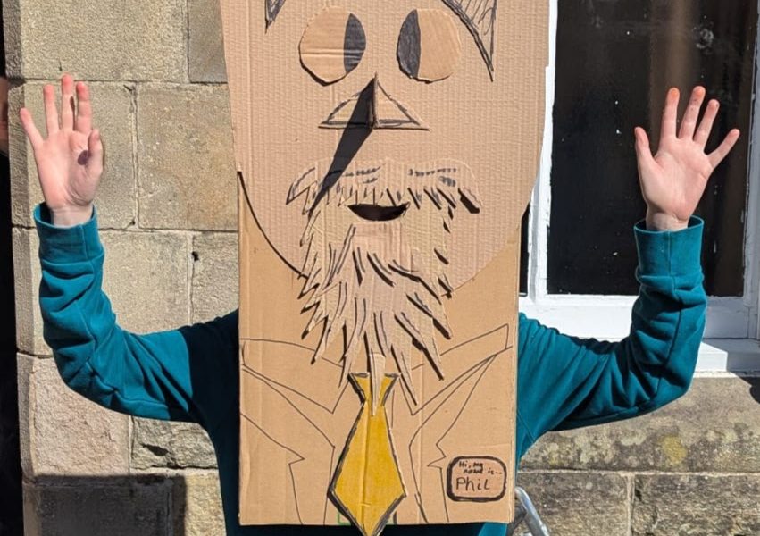 A person is wearing their cardboard creation over their head. It has been made from a large cardboard box and looks like a funny bearded character with a yellow tie
