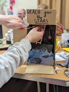 The image is a close up of someones hands adding detail to an art project that fits inside a small cardboard box. On top of the box is a slogan saying "Reach For The Stars"