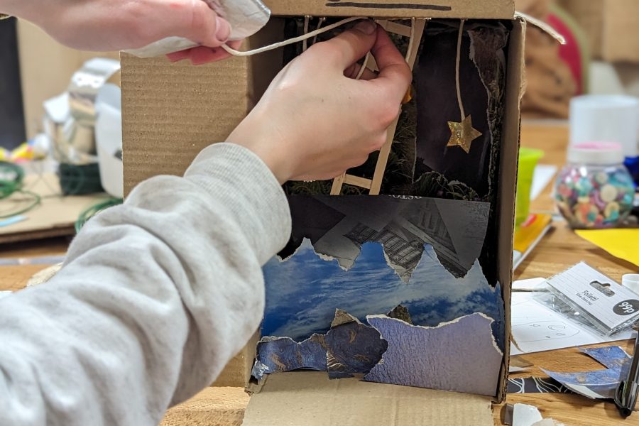 The image is a close up of someones hands adding detail to an art project that fits inside a small cardboard box. On top of the box is a slogan saying "Reach For The Stars"