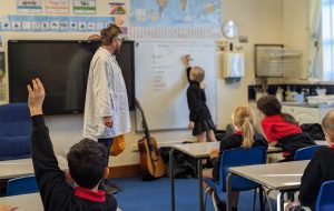 The photo is taken from the back of a classroom. Children are at their desks, one with their hand up. A girl is at the whiteboard writing something and an actor looks on and he is dressed in a white lab coat, pretending to be a scientist
