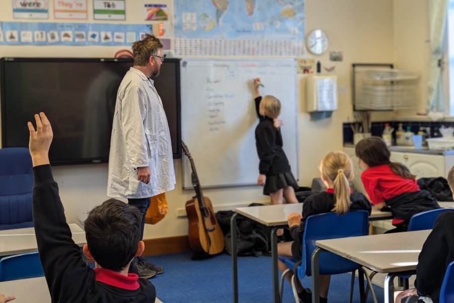 The photo is taken from the back of a classroom. Children are at their desks, one with their hand up. A girl is at the whiteboard writing something and an actor looks on and he is dressed in a white lab coat, pretending to be a scientist