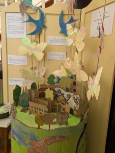 There is aa display on a table top of cardboard houses and Silk moths and swifts fly above it. The artwork was created by the community