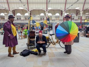 Our Curing Chemists dressed in Victoriana style costumes are having fun with a man who is sat on a stool and laughing. One of the actors is holfing a colourful umbrella with words written on it of various ailments and they are trying to predict what is wrong with him.