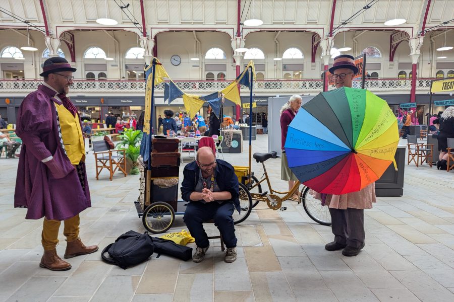 Our Curing Chemists dressed in Victoriana style costumes are having fun with a man who is sat on a stool and laughing. One of the actors is holfing a colourful umbrella with words written on it of various ailments and they are trying to predict what is wrong with him.