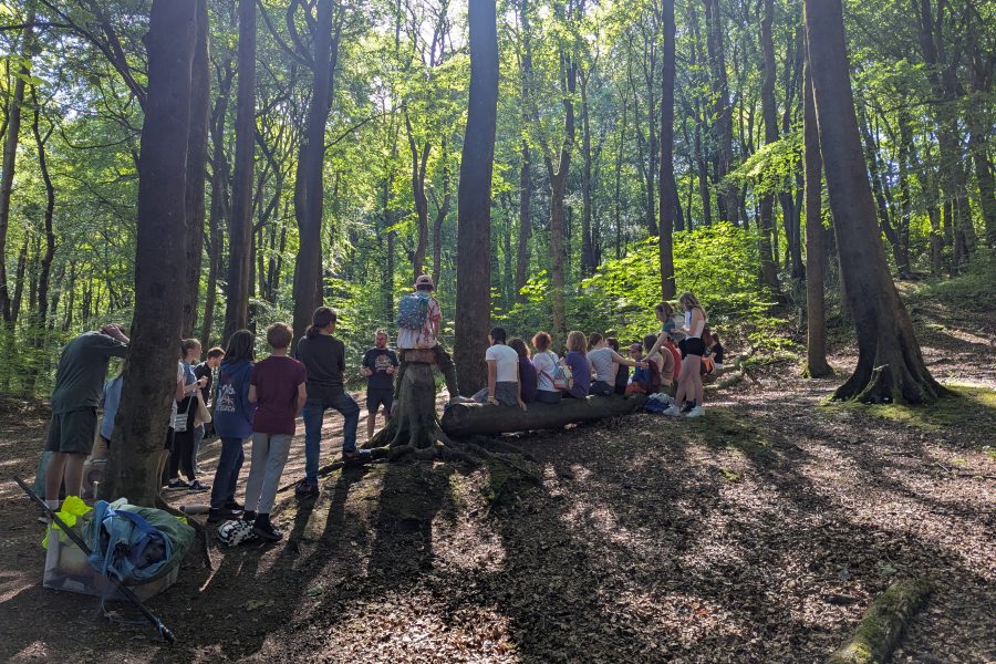 In A beautiful woodland with the sunlight dappled by the leaves, children gather round as their director gives them a team talk before a production.
