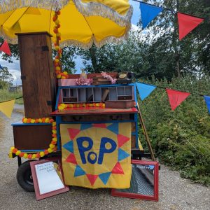 A brightly coloured cart filled with various pieces of coloured furniture stands outside. There are mirrors, bunting and flowers decorating it.