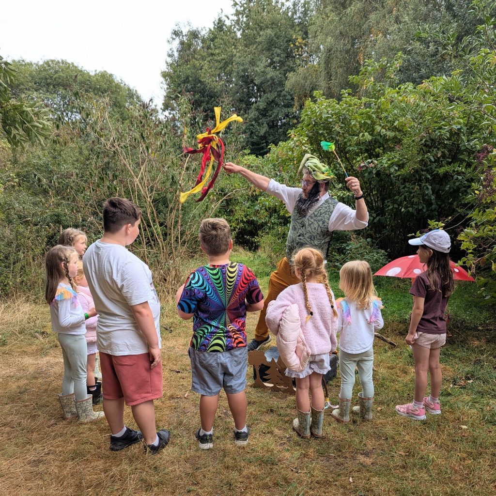 Children gather around a storyteller. They are outside in a clearing with green bushes surrounding them