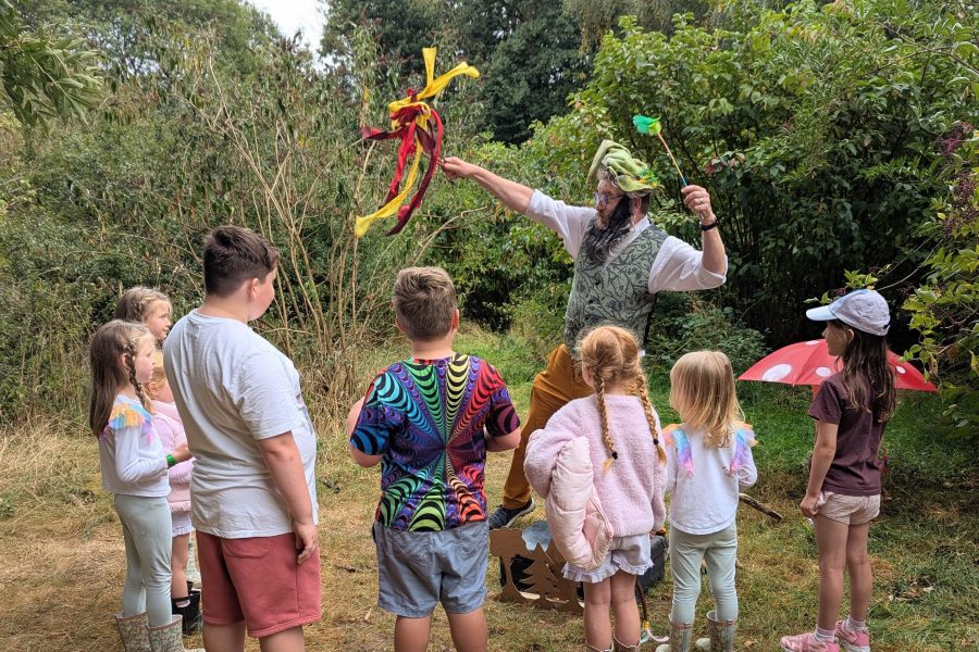 Children gather around a storyteller. They are outside in a clearing with green bushes surrounding them