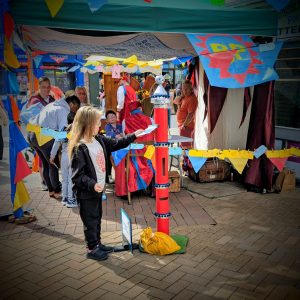 A child stands in front of a tall funny looking postbox which is red, silver and blue. There are flags decorating the space and a gazebo in the background. This is also decorated with bunting which is yellow, blue and red.
