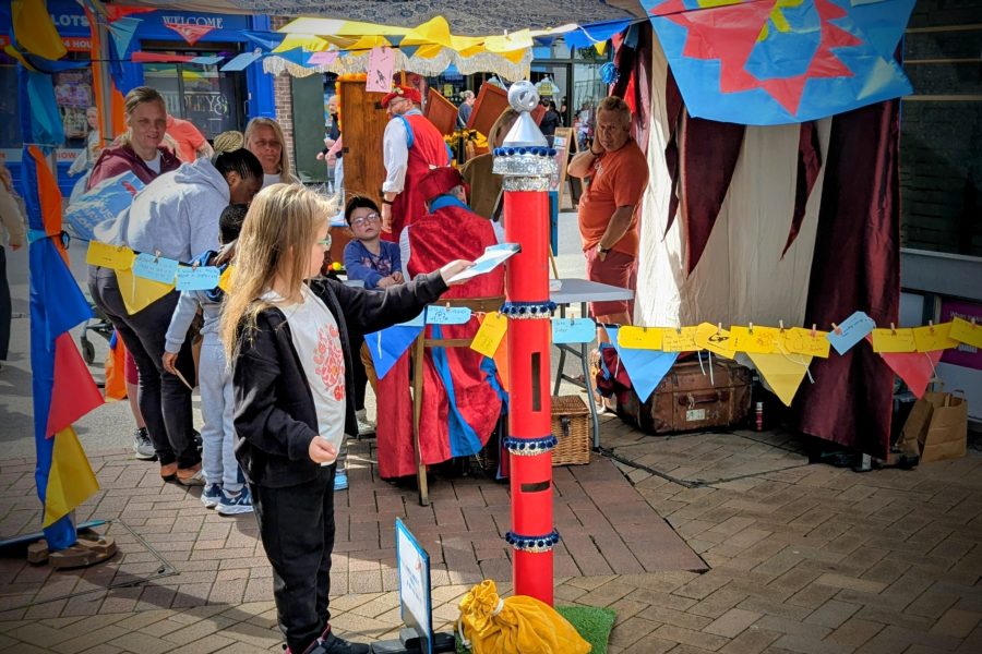 A child stands in front of a tall funny looking postbox which is red, silver and blue. There are flags decorating the space and a gazebo in the background. This is also decorated with bunting which is yellow, blue and red.