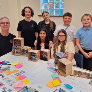 5 young people and two artists from Babbling Vagabonds sit and stand around a table covered in artwork and design ideas for the new commission of The Post Office Of Posibilities