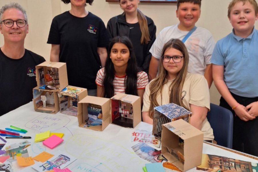 5 young people and two artists from Babbling Vagabonds sit and stand around a table covered in artwork and design ideas for the new commission of The Post Office Of Posibilities