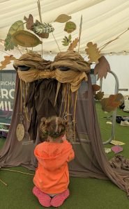 A small child with her back to the camer and in an orange top and plaits in her hair sits in front of our big puppet Pete. Pete has big eyes and brown fabric falls to form his body.