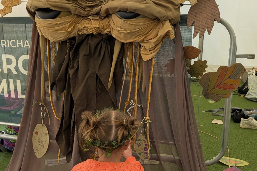 A small child with her back to the camer and in an orange top and plaits in her hair sits in front of our big puppet Pete. Pete has big eyes and brown fabric falls to form his body.