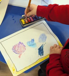A reception pupil paints dots on a page. You can see there painting and them using the palette of colours