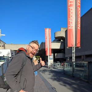 Phil, Artistic Director of Babbling Vagabonds is smiling with his thumb up outside The National Theatre. It is a sunny day with bright blue skies