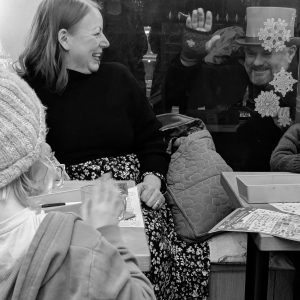 Black and white photo of a woman smiling looking out of a shop window, while on the otherside a man dressed in victorian clothes and a top hat is smiling and waving