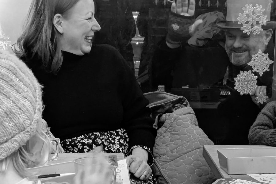 Black and white photo of a woman smiling looking out of a shop window, while on the otherside a man dressed in victorian clothes and a top hat is smiling and waving