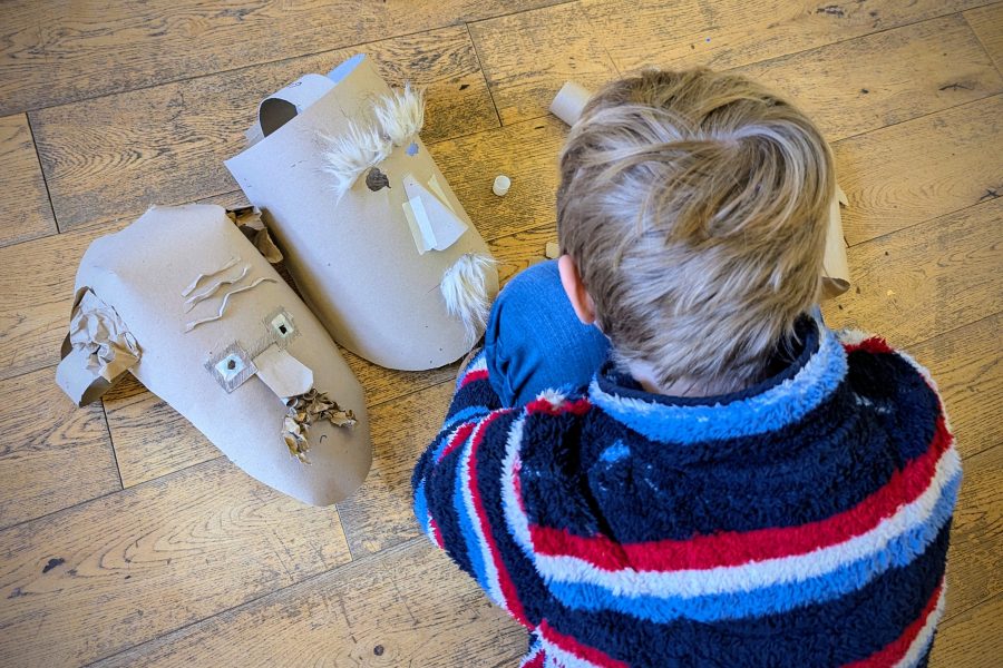 The photo is taken from above and a young boy sits on the wooden floor and makes a mask from cardboard.