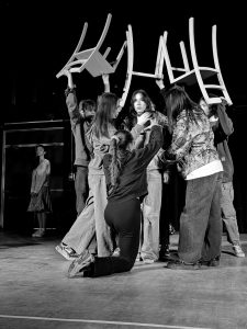 A black and white image of a concerned teenager being surrounded by other teenagers holding chairs above their heads in a threatening way