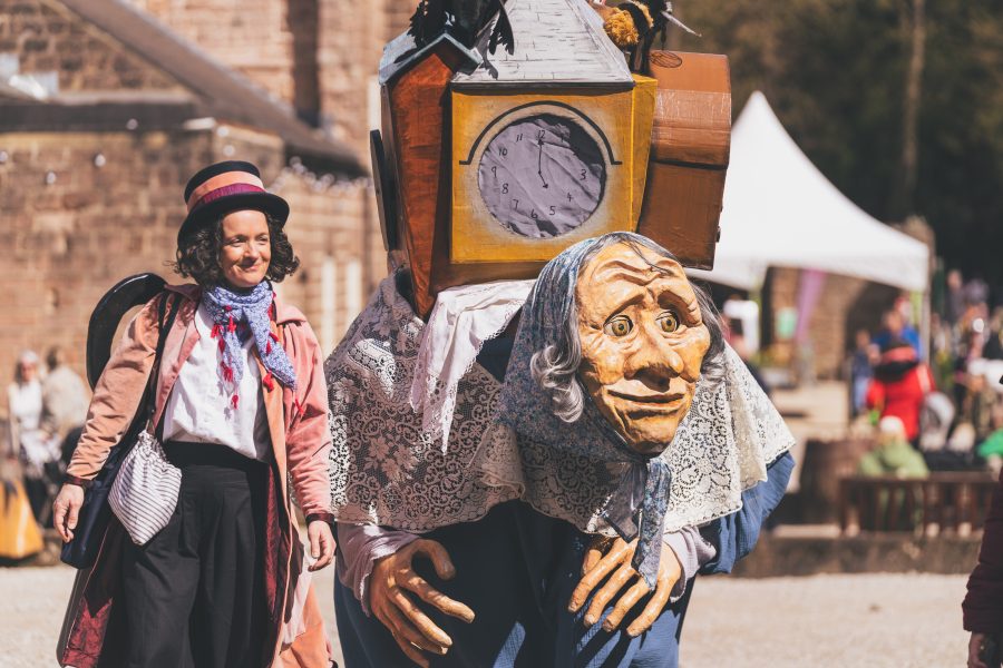 A large puppet designed llike an old lady carrying herlife on her back wanders cromford mill courtyard with their helper who is dressed in a pink coat and wearing a top hat