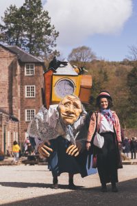 A large walk about puppet of an old lady that is carrying her life on her back. She walsk with a costume character of a woman in a pink coat and top hat