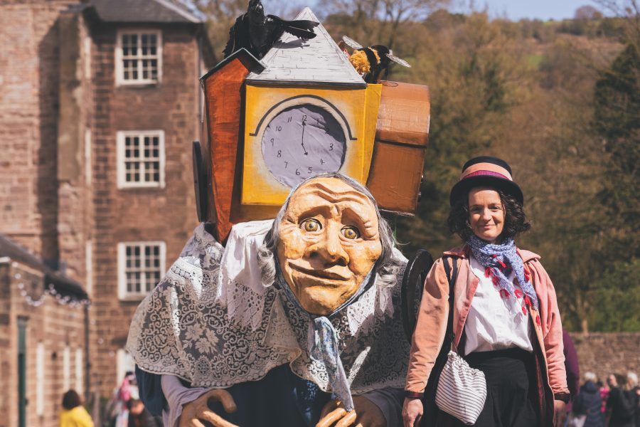 A large walk about puppet of an old lady that is carrying her life on her back. She walsk with a costume character of a woman in a pink coat and top hat