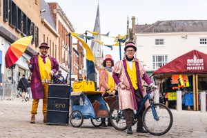 Three costumes characters stand proudly with a bicycle and trailer with yellow and blue flags on a market square. They are the curing chemist ready to dispense remedies to passer bys