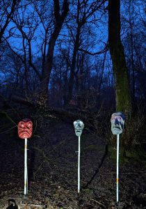 Three large gargoyle style faces on poles in the dark woods have been uplit and look out giving the photo and eerie feel.