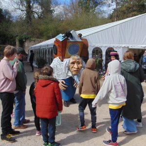 Children gather around a large walkabout puppet, that has been designed like an old lady carrying the world on her back