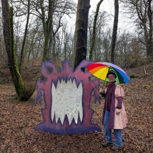 Mark is in the woods wearing a pink calf legth jacket and holding an umberella. He is standing next to a big pink and purple monster that is taller than him.