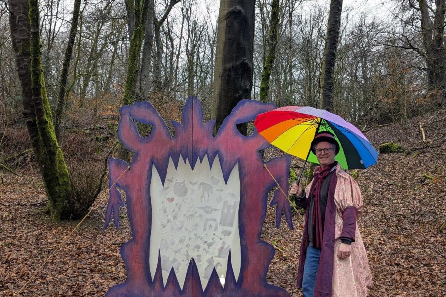 Mark is in the woods wearing a pink calf legth jacket and holding an umberella. He is standing next to a big pink and purple monster that is taller than him.