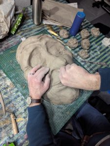 Hands sculpt a large head from clay. The head is in the style of an old lady
