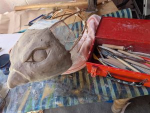 A clay head of a swift has been sculpted and sits on a willow framework which will eventually be the body. A red toolbox of clay tools is next to it on the table